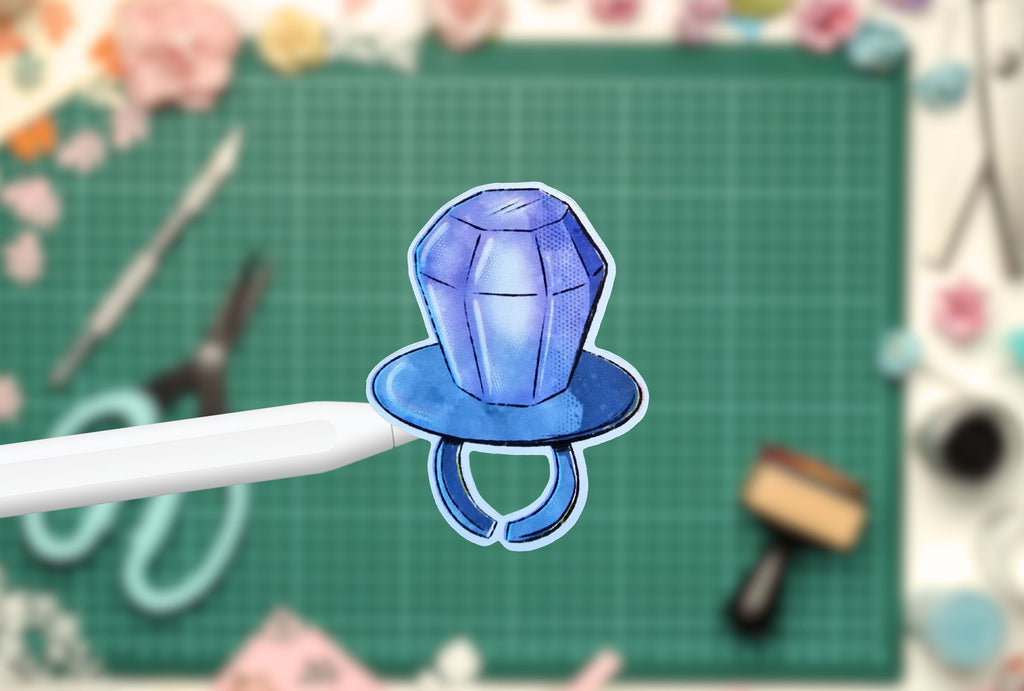 Blue gem-shaped candy sticker on a white pen against a green cutting mat with tools in the background