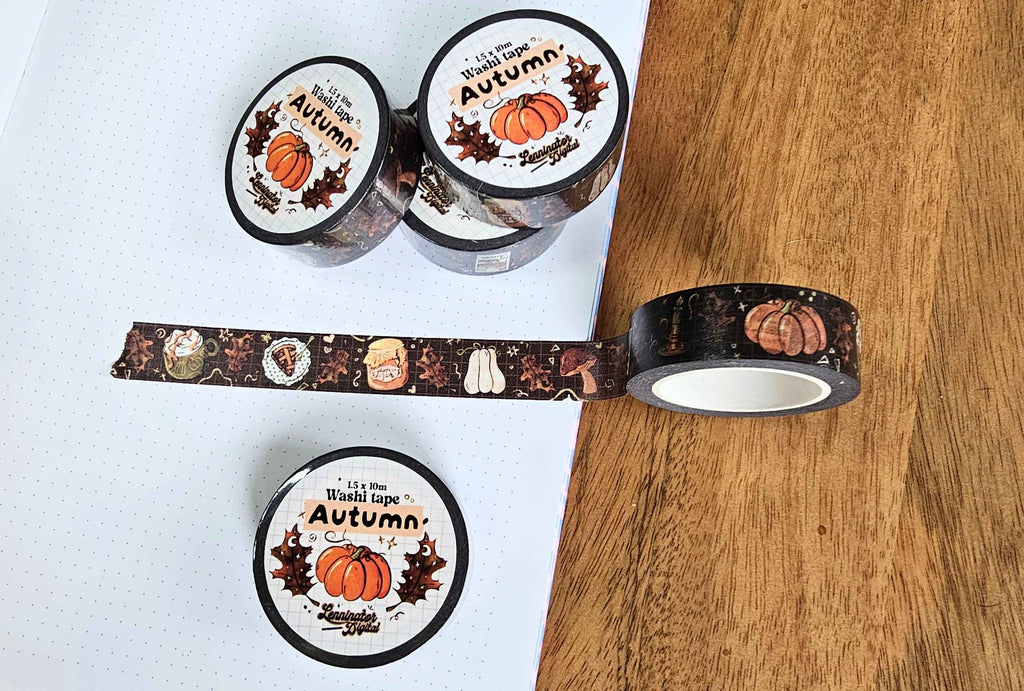 Three rolls of decorative washi tape with an autumn theme are displayed on a white dotted notebook and a wooden surface. The tape features illustrations of pumpkins, acorns, leaves, and the text 'It's Fall, So I'm Washi Tapes Autumn'.