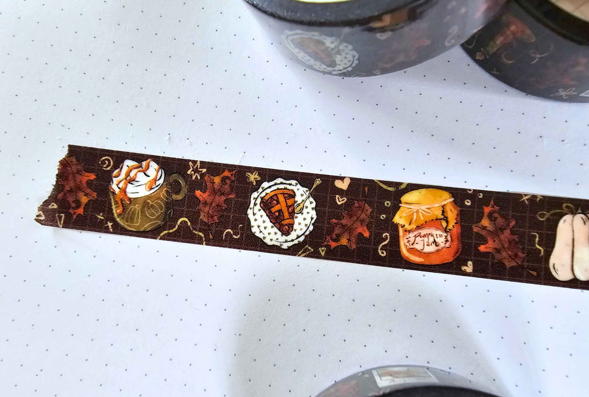 A close-up of a roll of decorative washi tape featuring a fall coffee theme, laid out on a white grid paper background. The tape has illustrations of a coffee mug with whipped cream, a coffee cup with a striped drink, a coffee cup with a heart, a coffee cup with a face, autumn leaves, and a coffee jar.