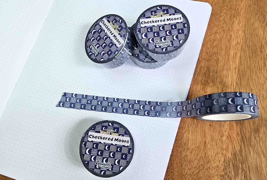 Three rolls of washi tape with a dark blue and gray 'Checkered Moons' celestial pattern, displayed on a white dotted notebook next to a wooden surface.
