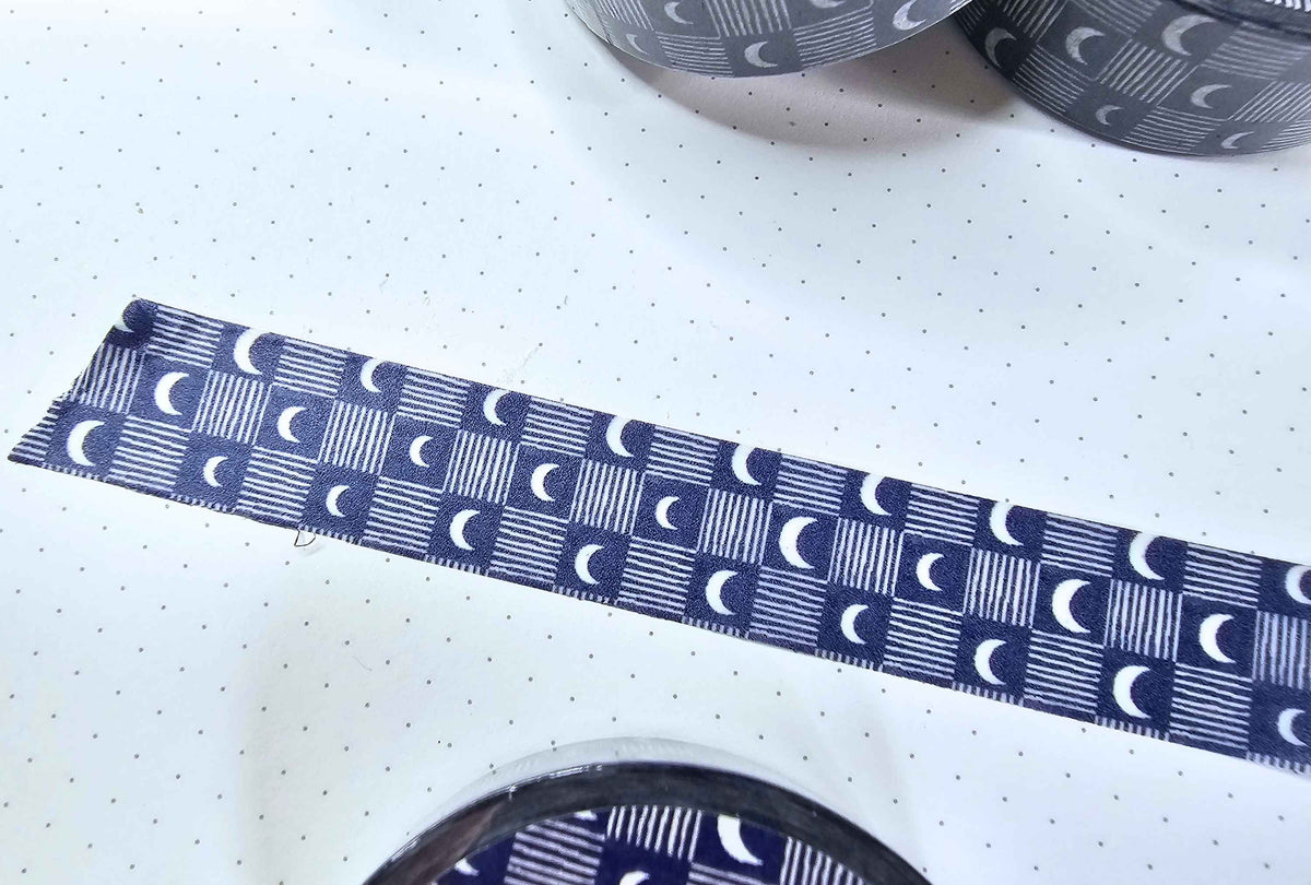 A close-up of a roll of patterned tape unrolled across a white surface. The tape has a repeating design of white crescent moons and vertical stripes on a dark navy blue background.