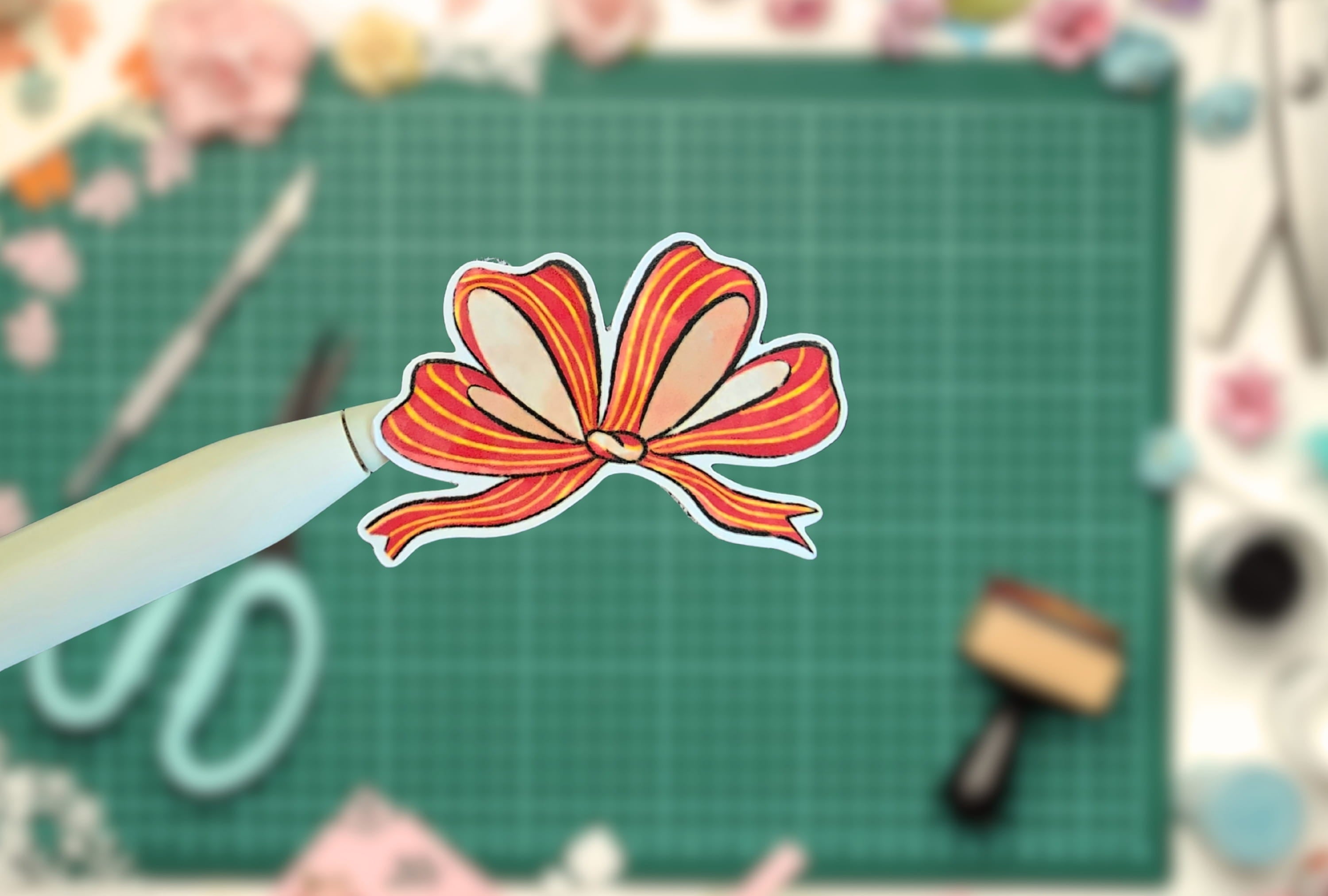 Colorful paper bow on a white pen against a green cutting mat with crafting tools.