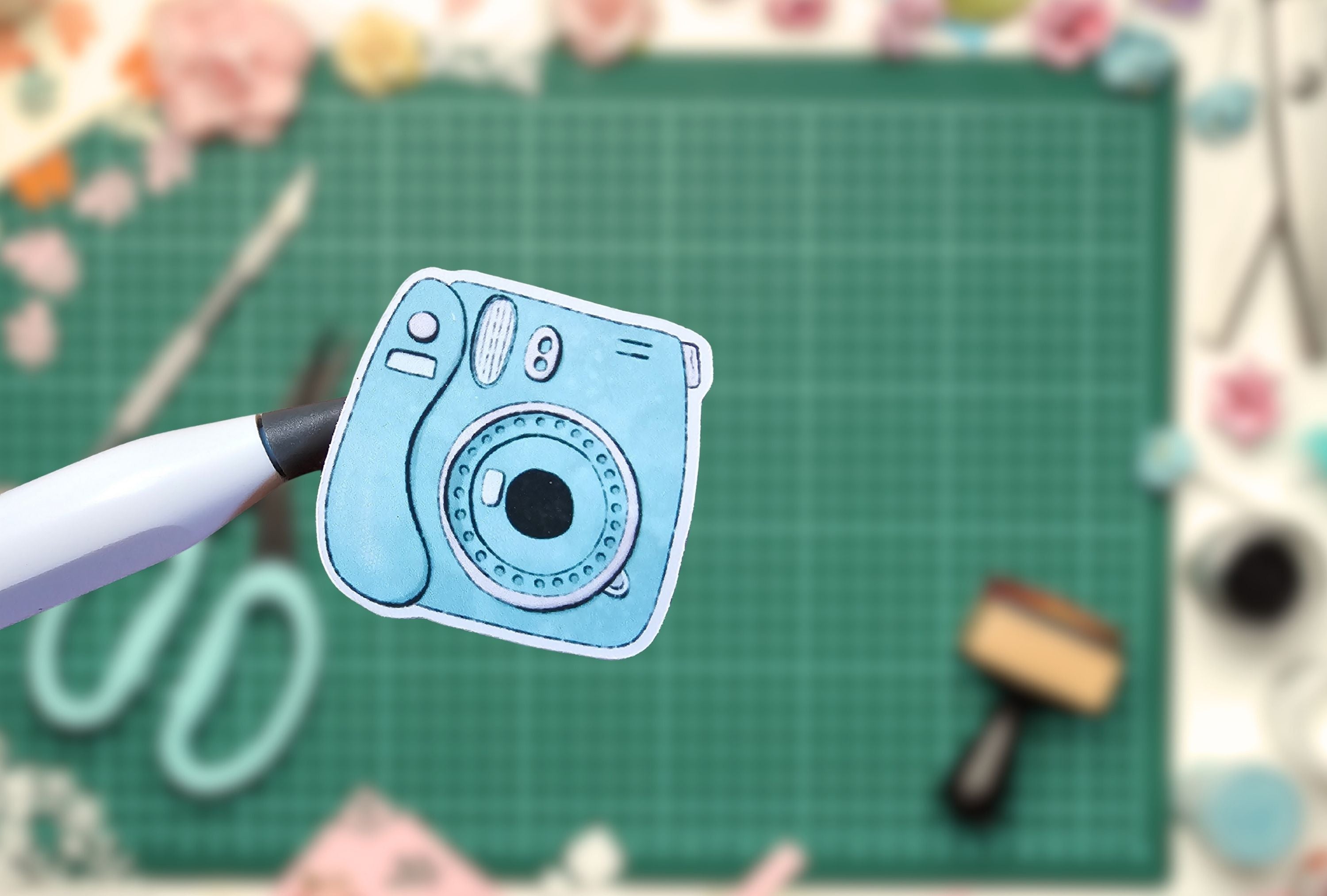 Blue camera-shaped sticker on a green cutting mat with crafting tools.