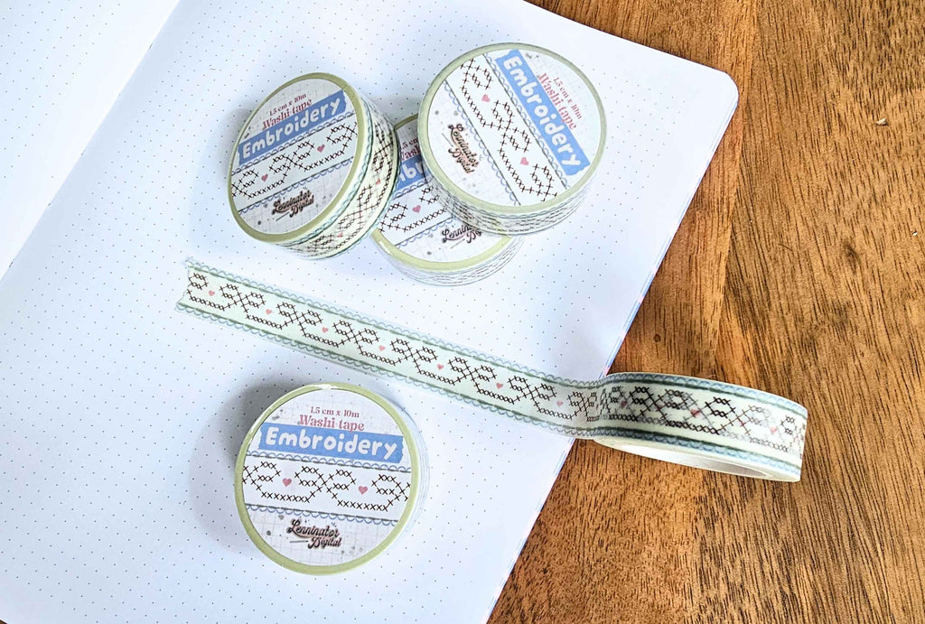 Embroidery washi tape rolls with a cross-stitch style pattern, displayed on a white notebook next to a wooden surface.