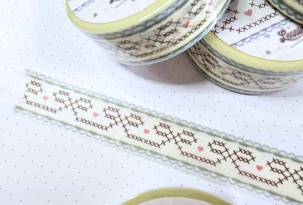A close-up of decorative washi tape with a cross-stitch style pattern in brown, light green, and small pink hearts, shown both on a strip and partially wound around rolls on a white dotted background.
