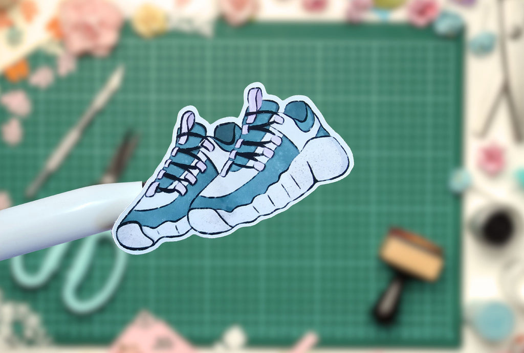 Cartoon-style blue sneakers held by a hand on a green cutting mat with tools in the background.