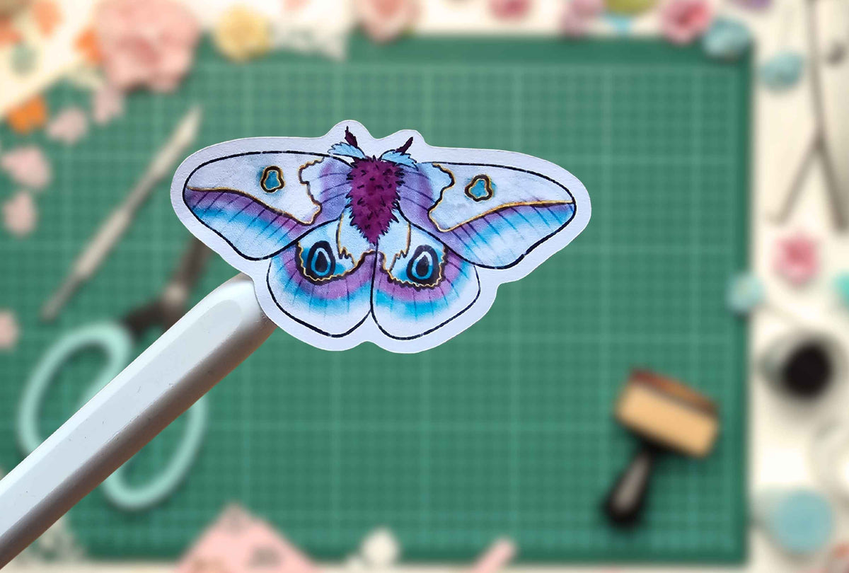 A die-cut sticker of a detailed painted moth with blue, purple, and white wings, held on a white stick against a blurred green cutting mat background.