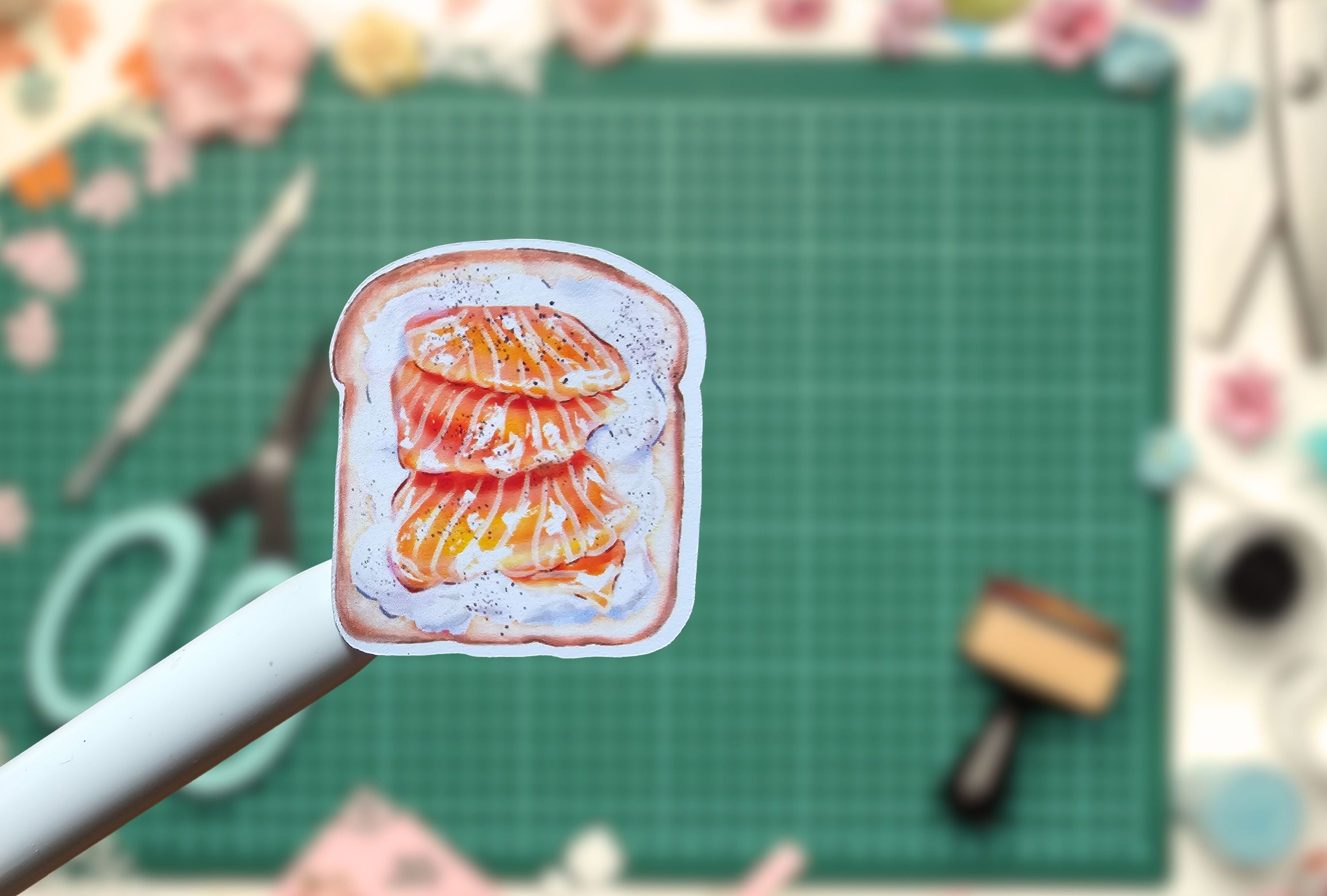 Sticker of a piece of bread with orange salmon on a white background, held by a white pen.