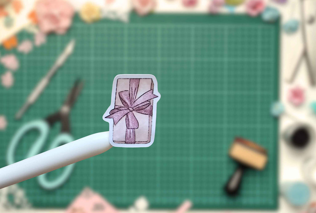 A small die-cut sticker of a wrapped gift box with a purple ribbon, held on a white stick against a blurred green cutting mat background with craft supplies.