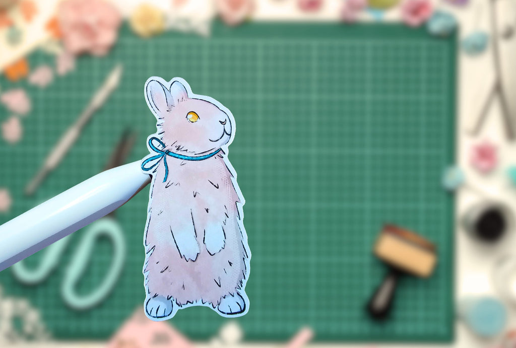 Cartoon rabbit sticker held by a pen on a green cutting mat with crafting tools.