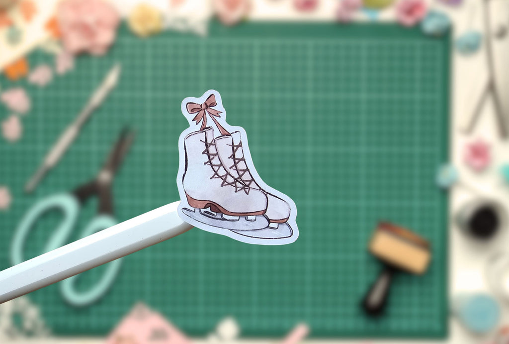A die-cut sticker of a white ice skate with brown laces and a pink bow, held on a white stick against a green cutting mat background with craft supplies.