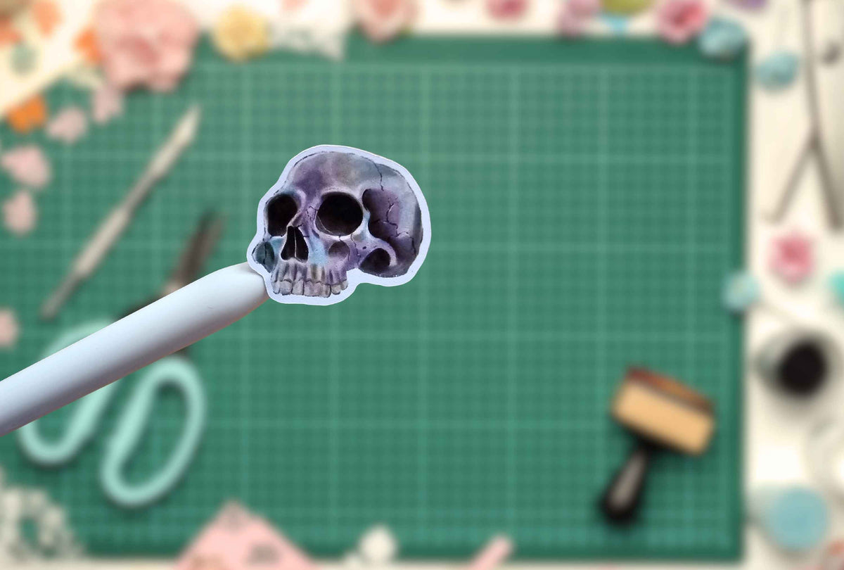 A small skull-shaped sticker or decal held on a white stick, with a green cutting mat and craft supplies blurred in the background.