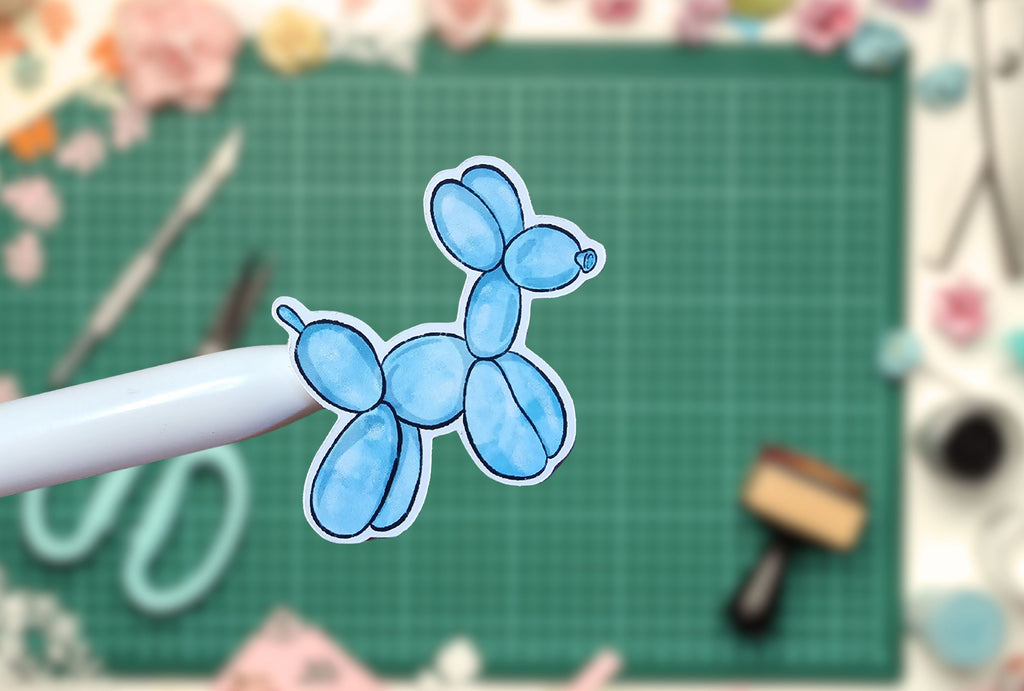 Blue balloon dog sticker on a white stick with a green cutting mat and crafting tools in the background.