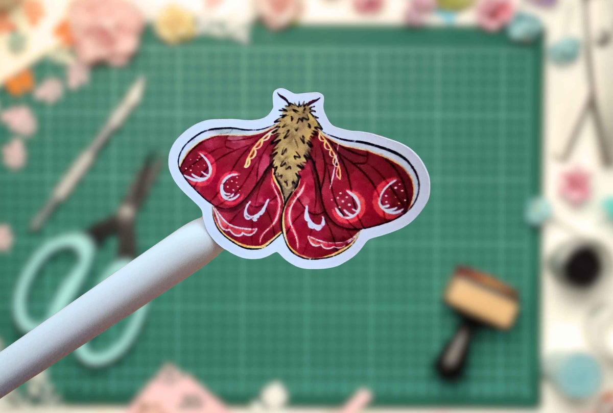 A die-cut sticker of a red and gold moth on a white stick, held up against a blurred green cutting mat background with craft supplies.