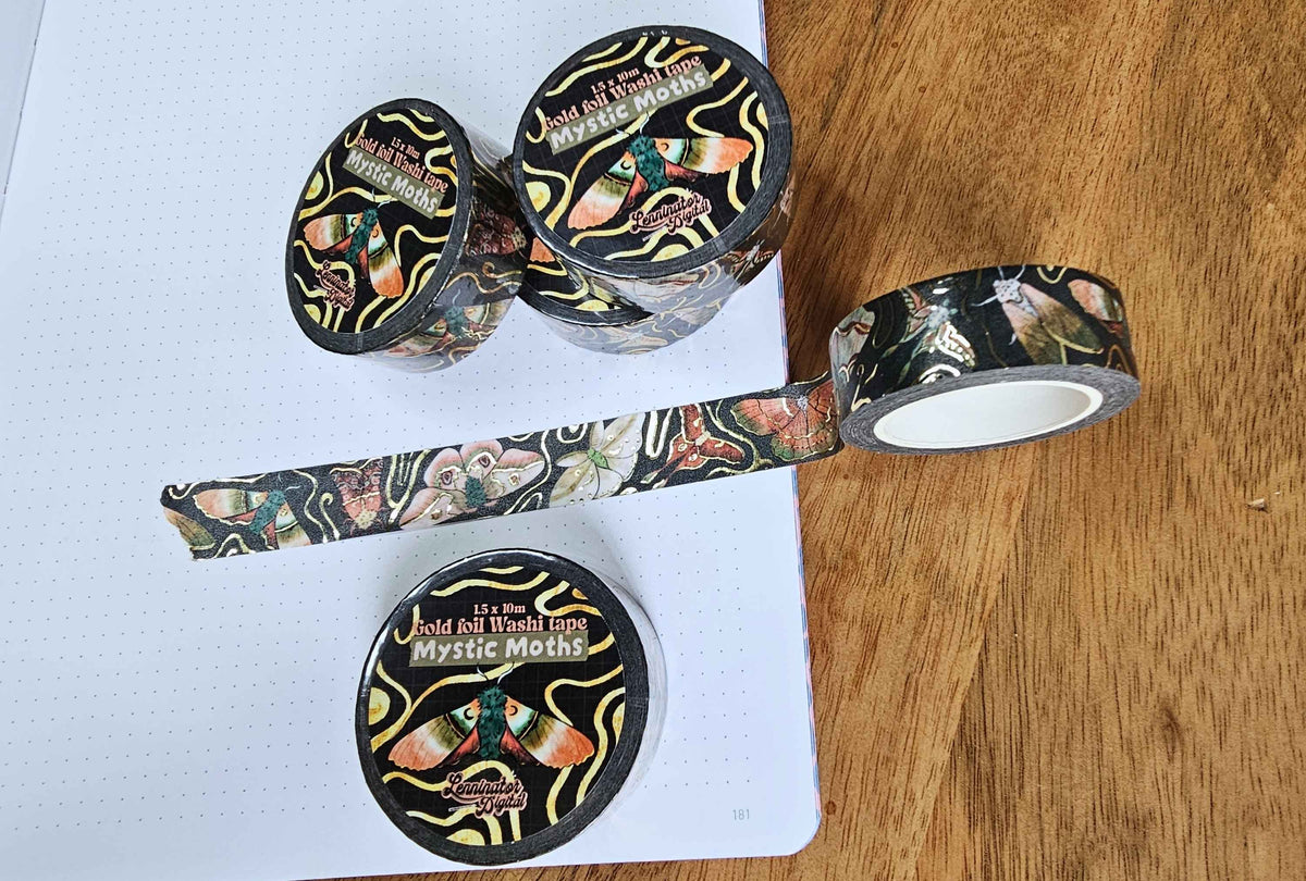 Three rolls of decorative washi tape with a black background and a colorful tattoo-style butterfly design, displayed on a white grid notebook and a wooden surface. 