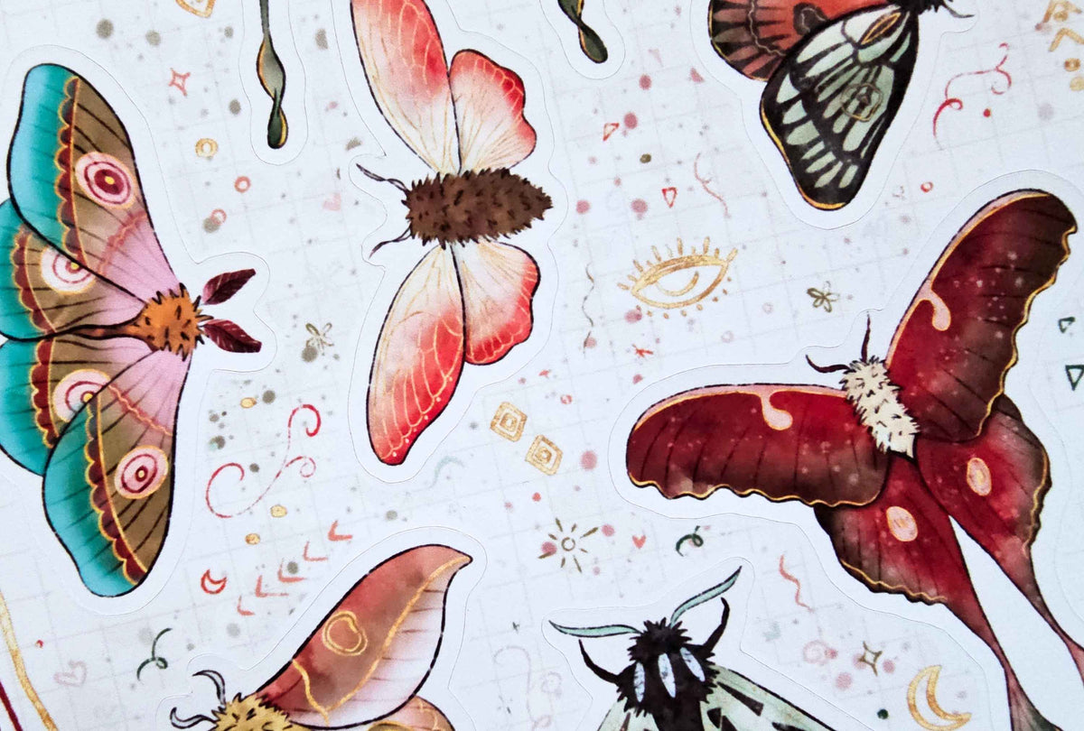 A close-up of a white paper surface decorated with watercolor-style illustrations of moths and butterflies in pink, red, teal, black, and gold accents.