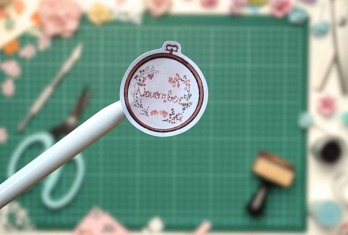 A small embroidered patch sticker with a floral design and the word 'november' is held on a white stick against a green cutting mat background with craft tools.