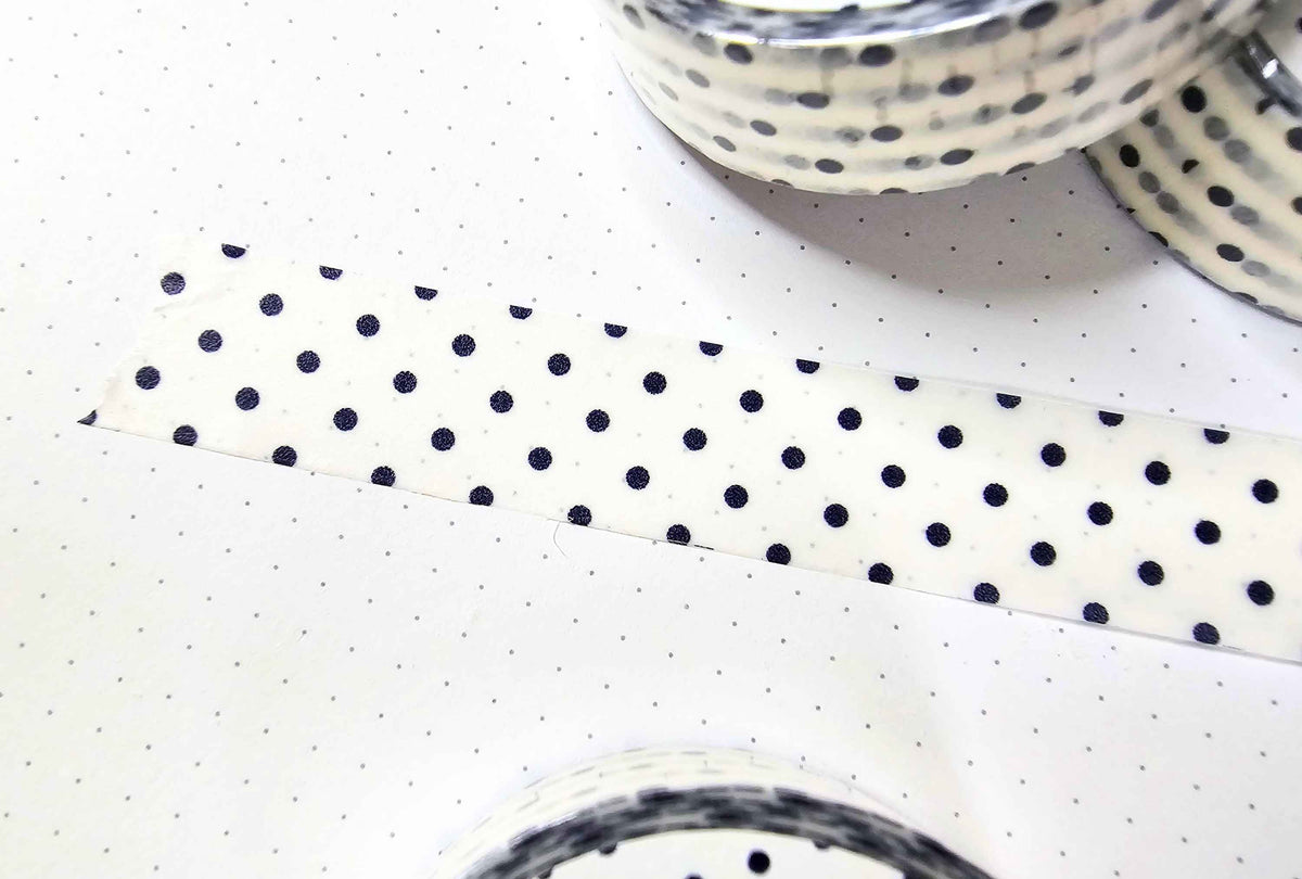 A close-up of a roll of washi tape with a navy blue polka dot pattern on a white background, laid diagonally across a white polka-dotted surface with other rolls partially visible.