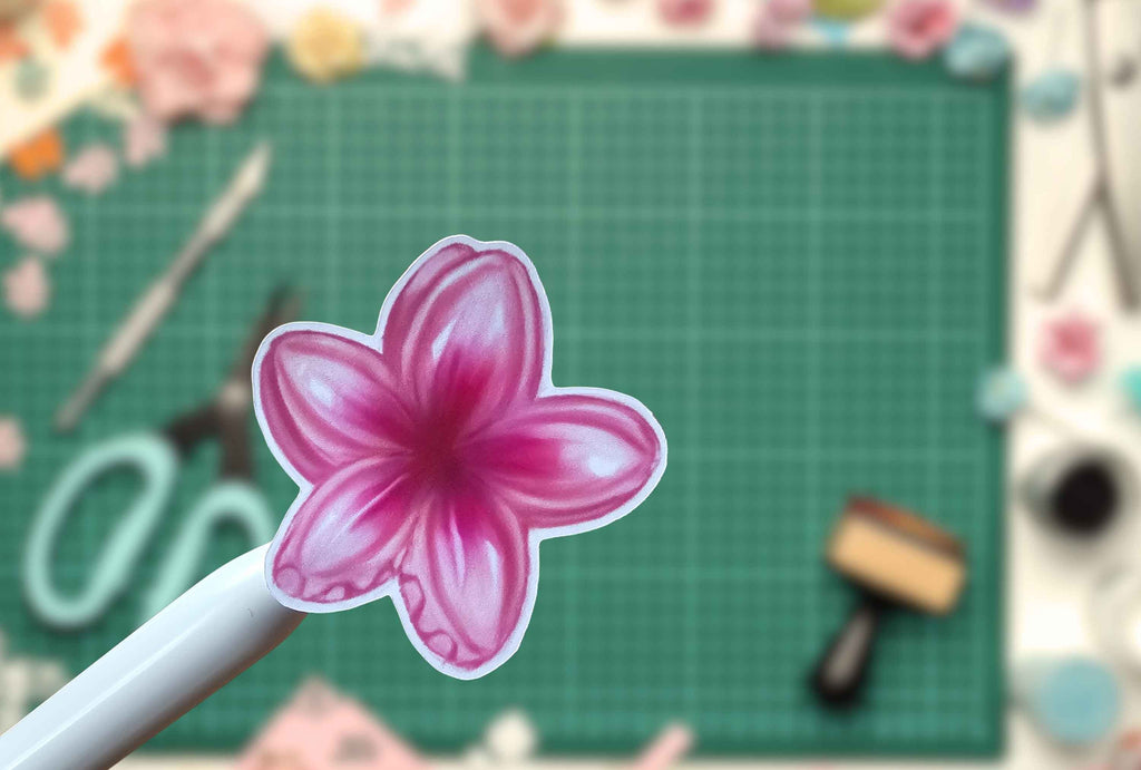 A pink flower-shaped sticker  held on a white stick, with a green cutting mat and craft tools blurred in the background.