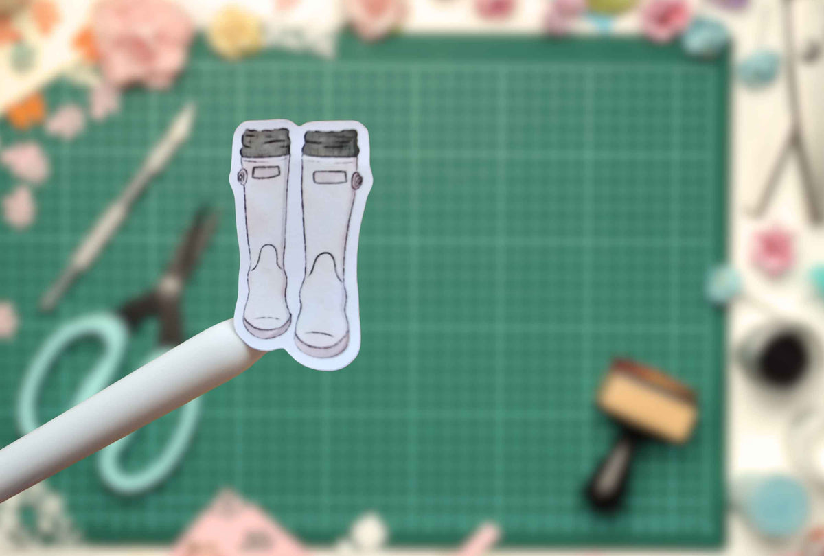 A small printed sticker showing a pair of white rain boots with dark gray cuffs, held on a white stick against a blurred green craft mat background with scissors, flowers, and a stamp visible.