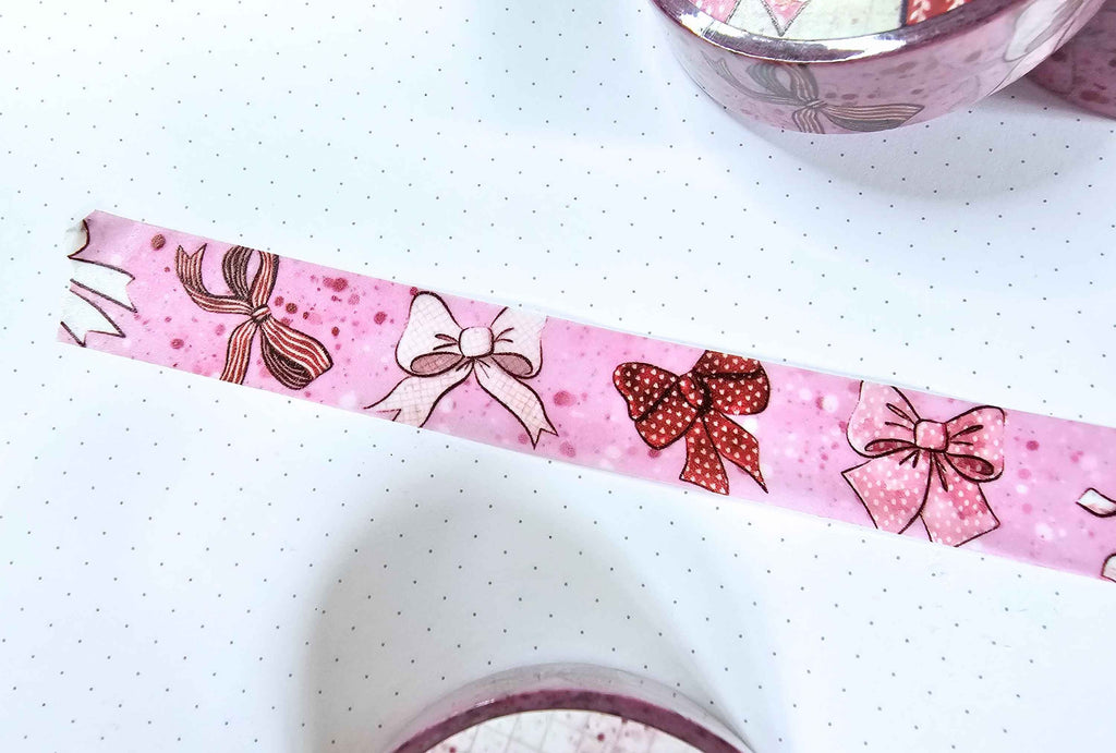 Pink washi tape with a bow pattern, shown partially unrolled on a white polka-dotted surface. The tape features various bows in brown, red, and white with pink glittery and solid details.