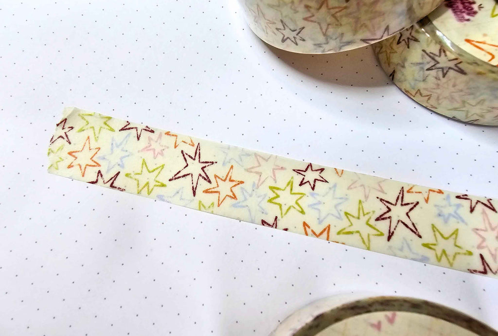 A close-up of a roll of washi tape with a colorful star pattern on a white background, with other rolls partially visible in the top right corner.
