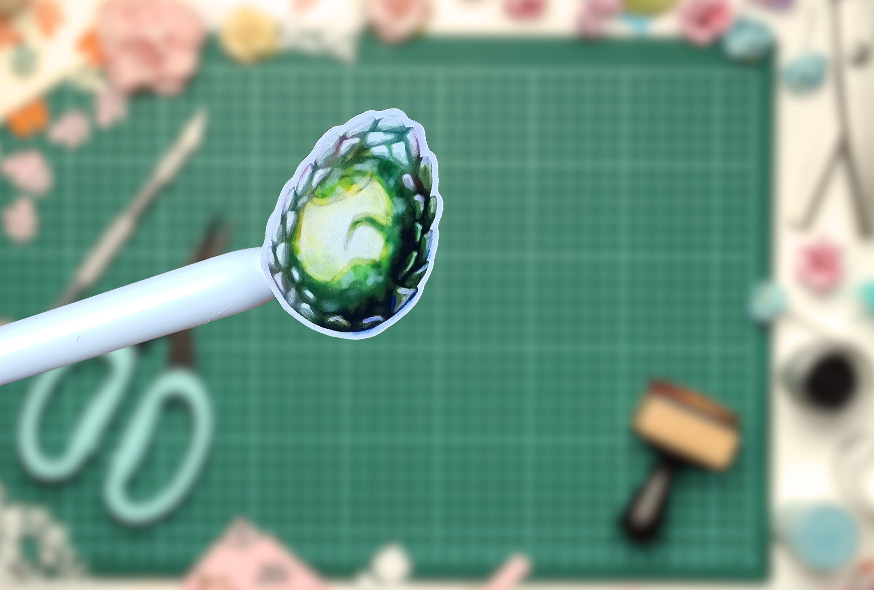 A close-up of a white-handled tool holding a small green dragon egg sticker, set against a blurred green cutting mat and craft supplies.
