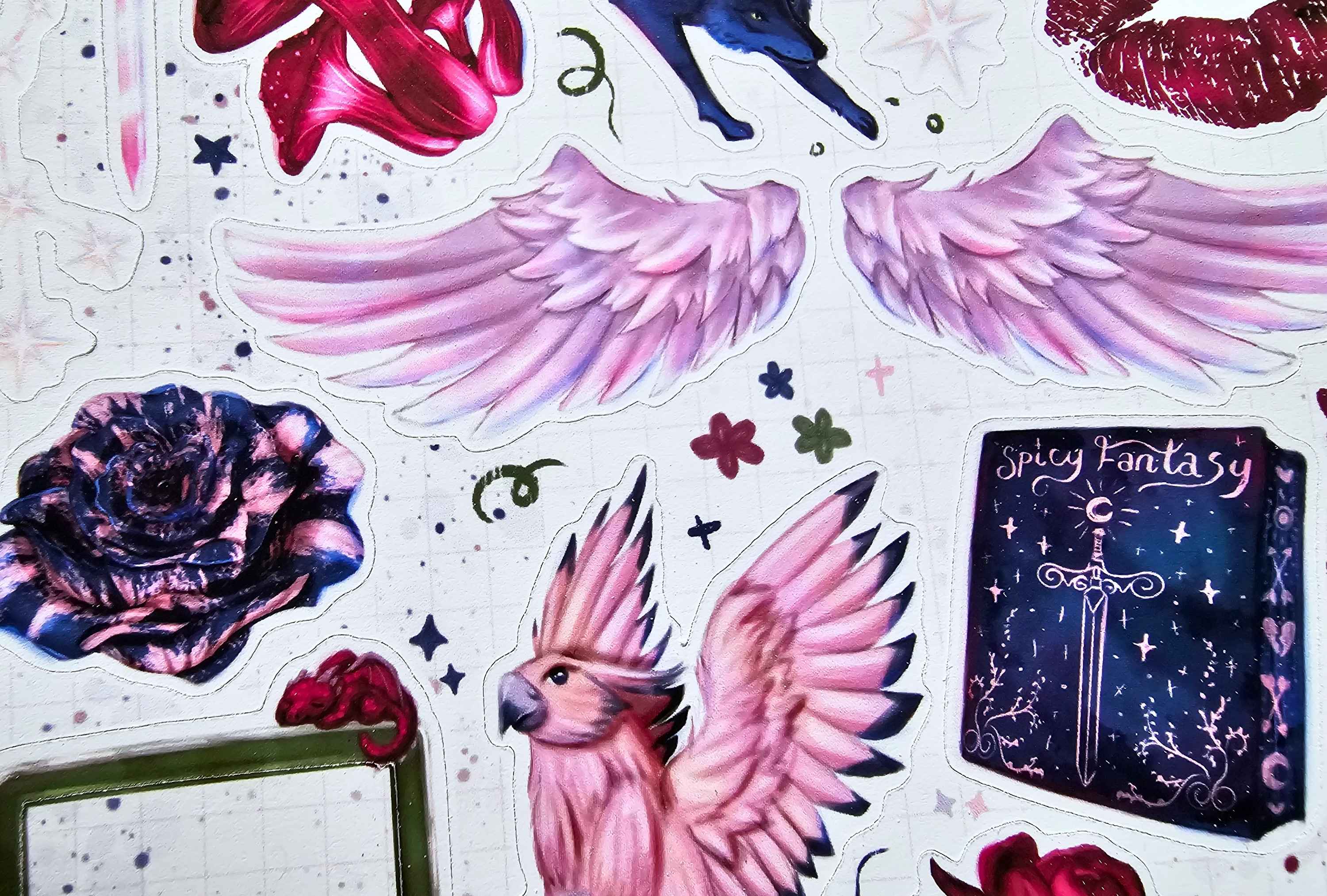 A sheet of decorative stickers featuring fantasy illustrations, including pink angel wings, a pink bird with wings, a dark blue book labeled 'Spicy Fantasy', a dark blue rose, and other small fantasy-themed accents on a white background.