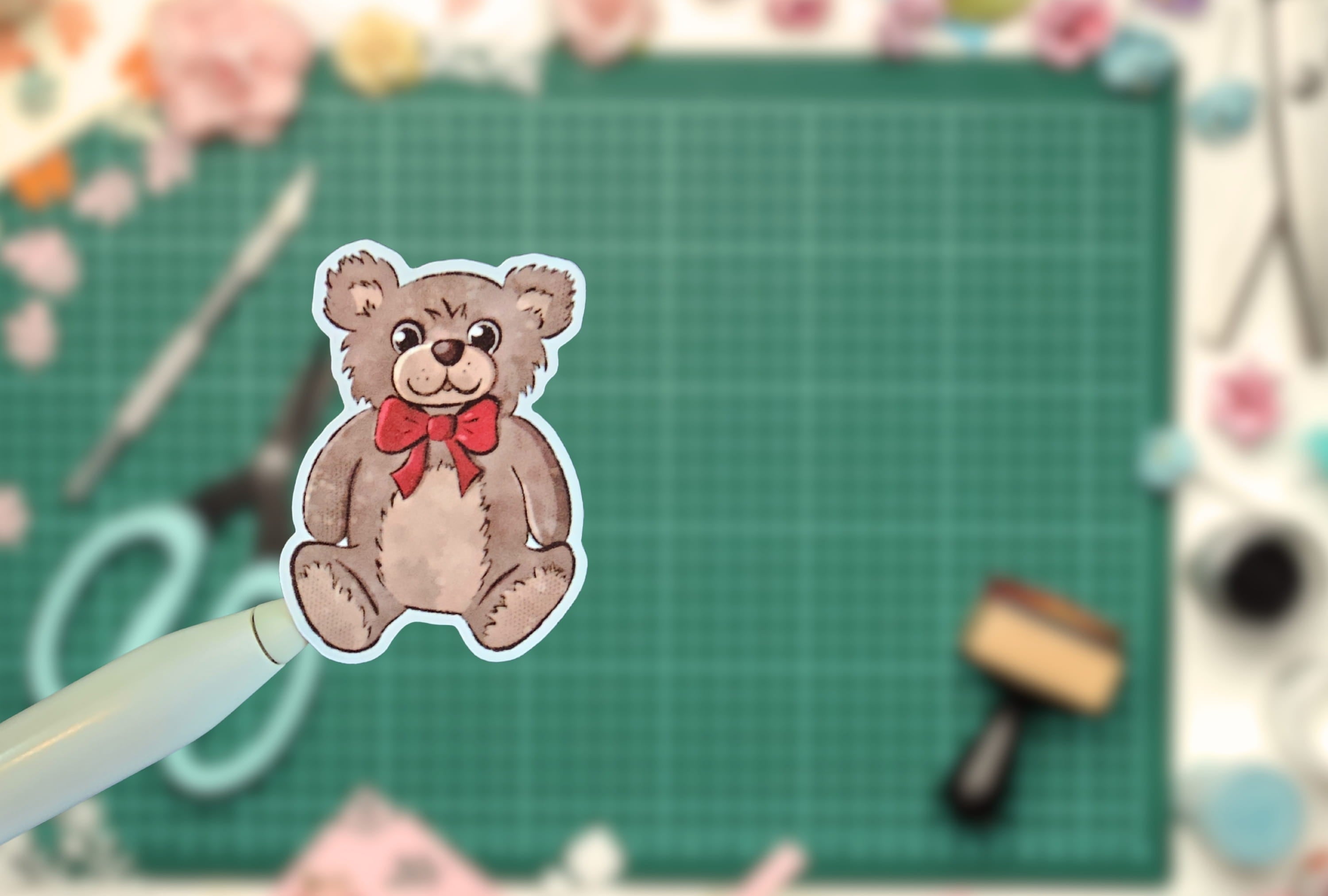 Sticker of a teddy bear with a red bow on a green cutting mat with crafting tools.