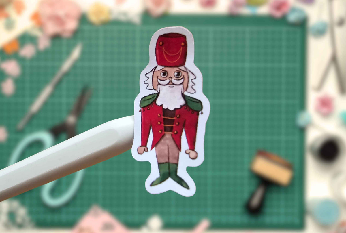 A die-cut sticker of a cartoon nutcracker soldier held by a white stylus, set against a blurred green cutting mat background with craft supplies.