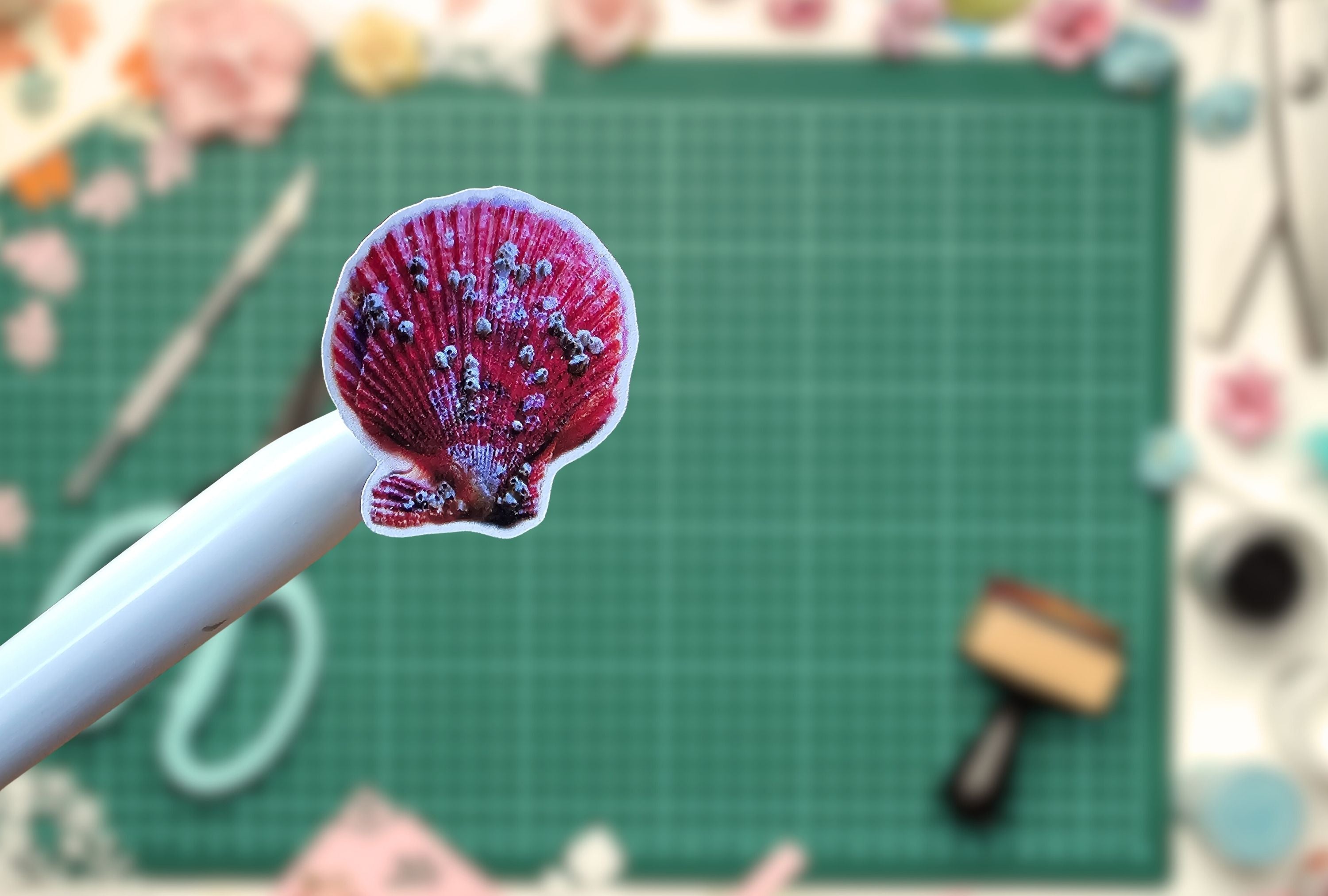 A small scallop shell-shaped sticker or decal with a pink and purple patterned design is held on a white tool over a green cutting mat with craft supplies blurred in the background.