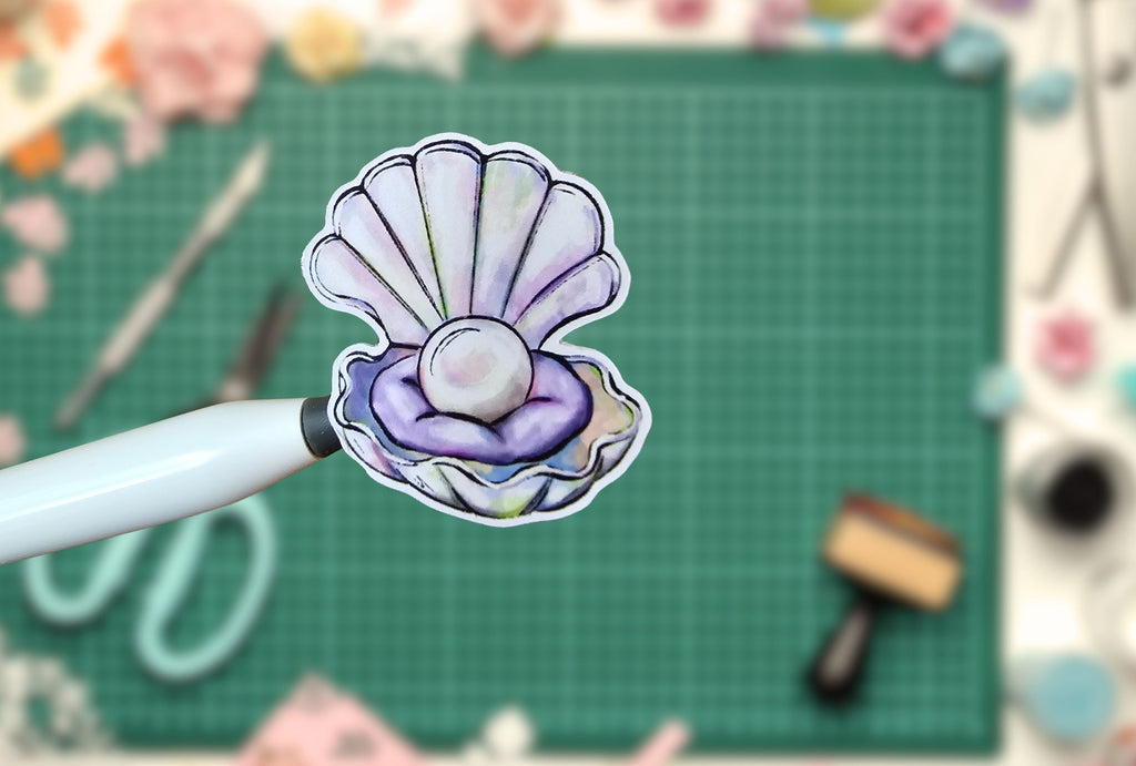 Stylized pearl in a shell sticker on a white pen against a green cutting mat with crafting tools.