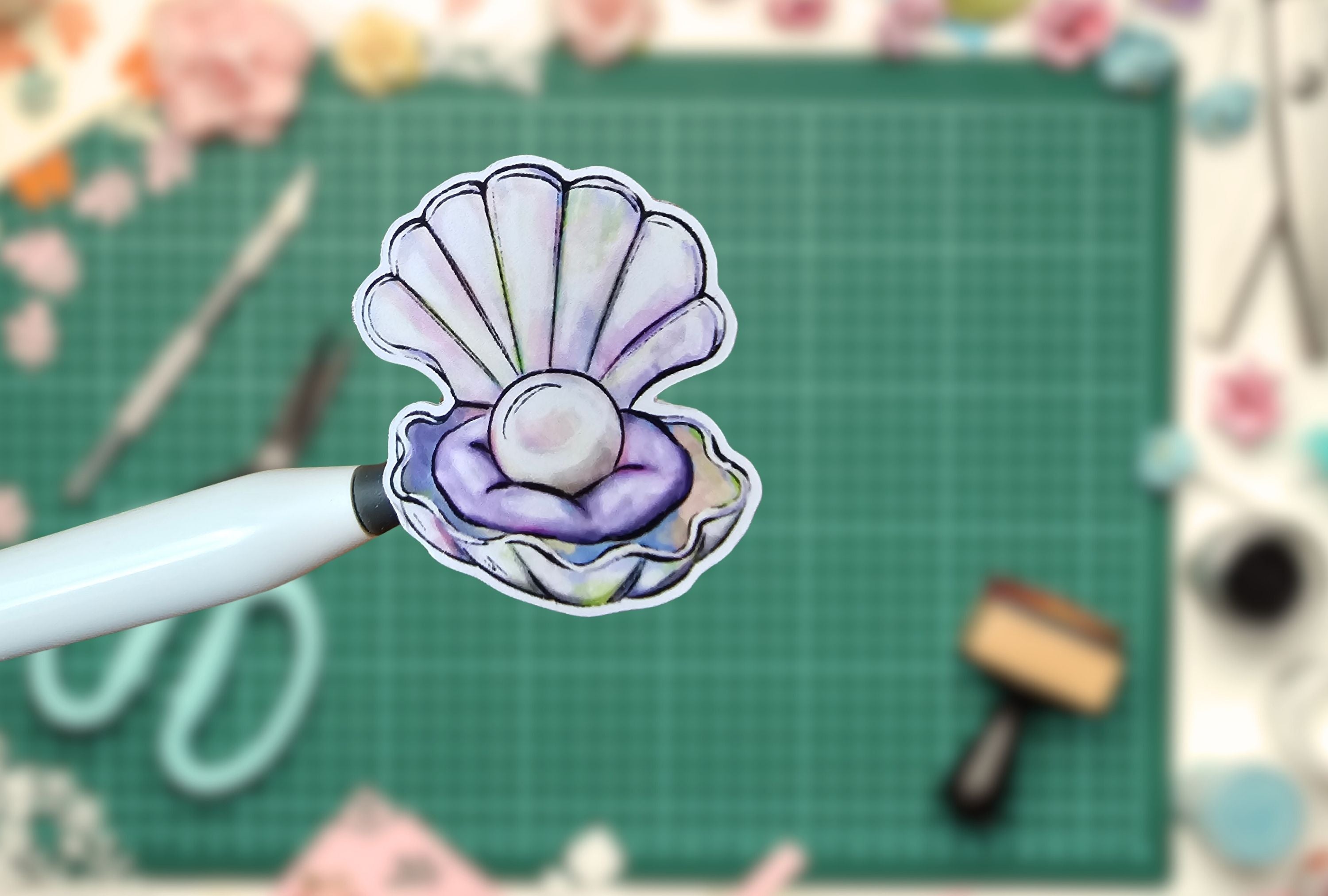 Stylized pearl in a shell sticker on a white pen against a green cutting mat with crafting tools.
