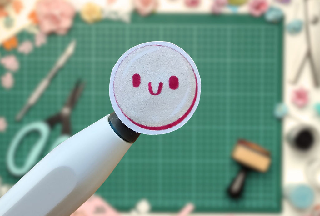 White pen with a circular sticker featuring a smiley face design on a green cutting mat with tools.