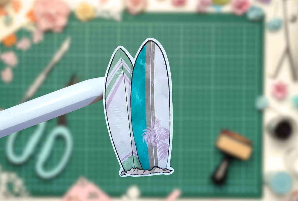 Decorative surfboard-shaped sticker on a cutting mat with crafting tools in the background