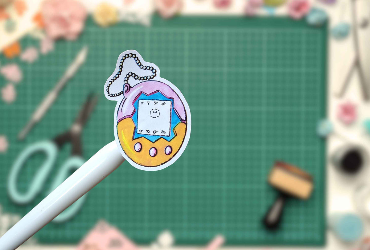 A hand-drawn Tamagotchi sticker held on a white stick, set against a blurred craft workspace with a green cutting mat and scissors.