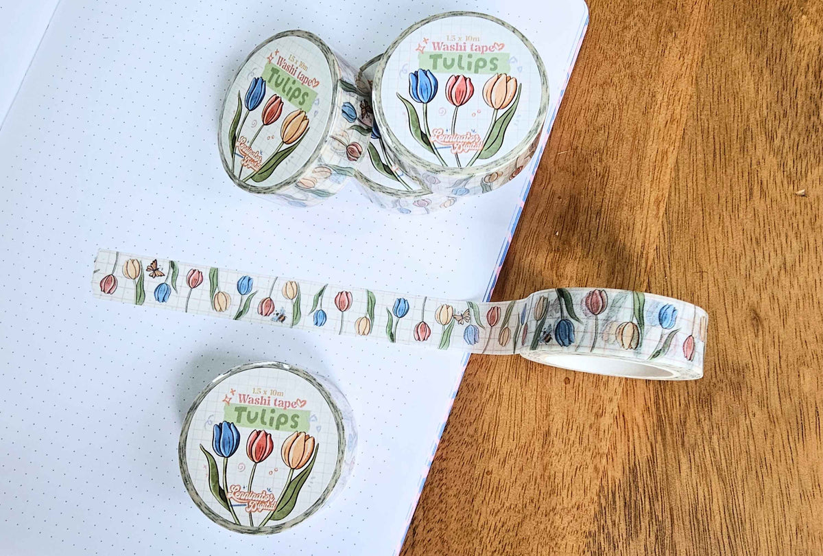 Three rolls of washi tape with a colorful tulip pattern are arranged on a white notebook and a wooden surface. The tape features illustrations of red, blue, and orange tulips with green leaves on a light background, and the top roll shows the brand label 'Washi Tape' and 'Tulips'.