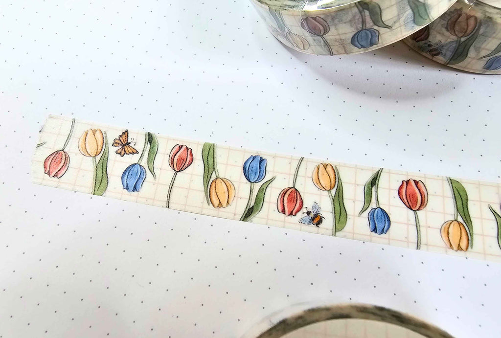 A close-up of a roll of washi tape featuring a pattern of colorful tulips, a butterfly, and a bee on a light grid background, with other rolls partially visible in the background.