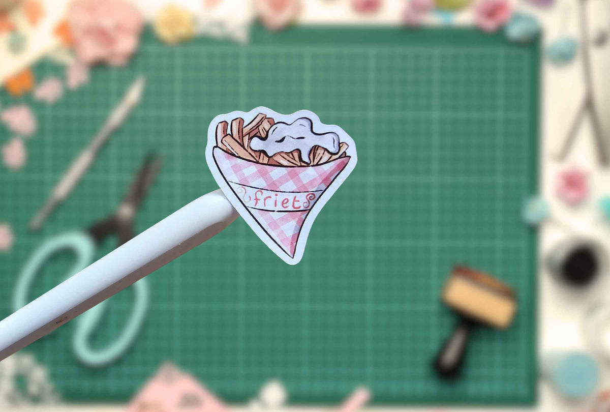 A small die-cut sticker of a pink gingham cone of fries with the word 'fries' written on it, held by a white stick against a green cutting mat background with blurred craft supplies.