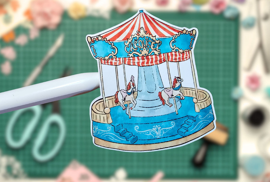 Paper cutout of a carousel with horses on a green cutting mat with scissors in the background