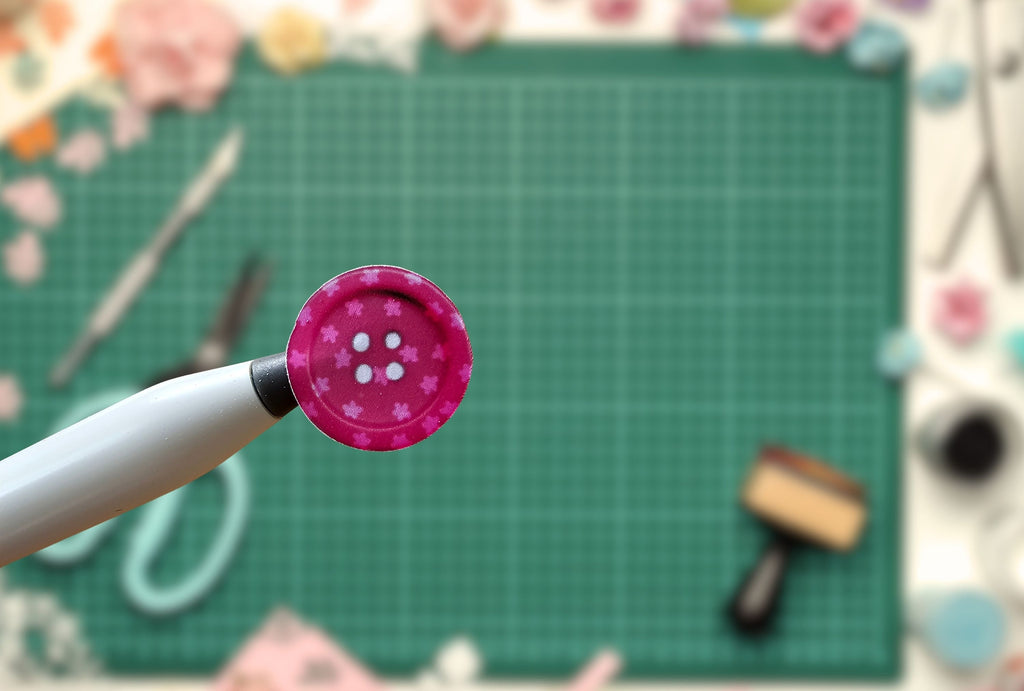 Pink button on a white handle against a blurred craft materials background