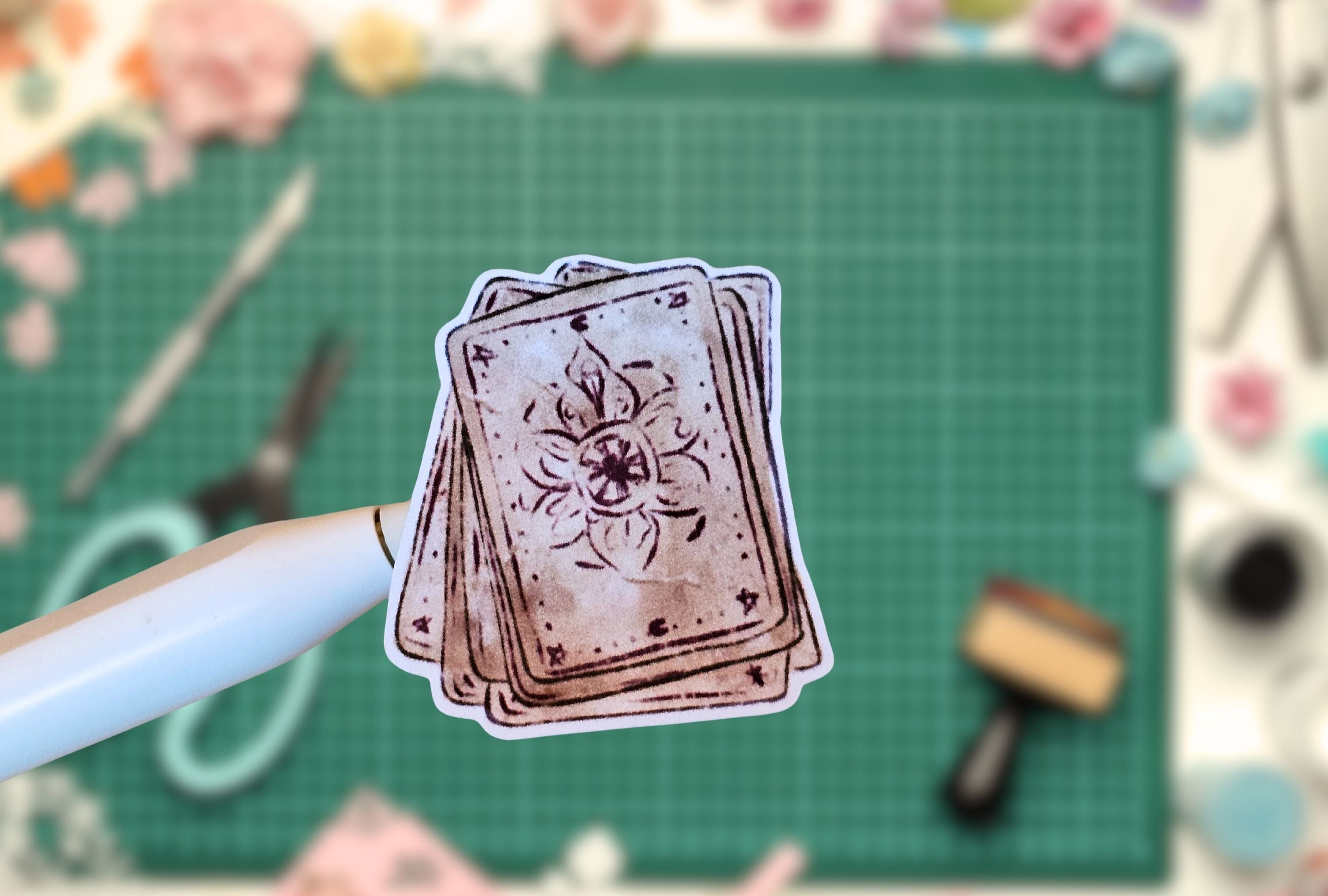 Stack of tarot cards held by a pen on a green cutting mat with crafting tools.