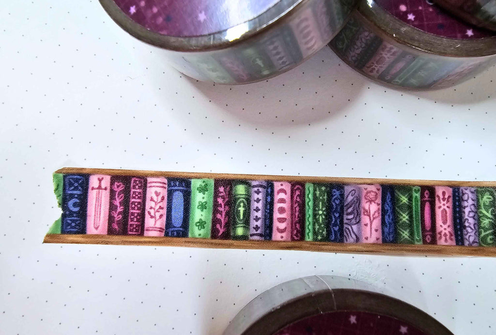 A close-up of a decorative washi tape roll laid flat, showing a row of colorful illustrated book spines in pink, green, purple, blue, and gold. Partial rolls of matching patterned tape are visible at the top and bottom on a white pegboard background.