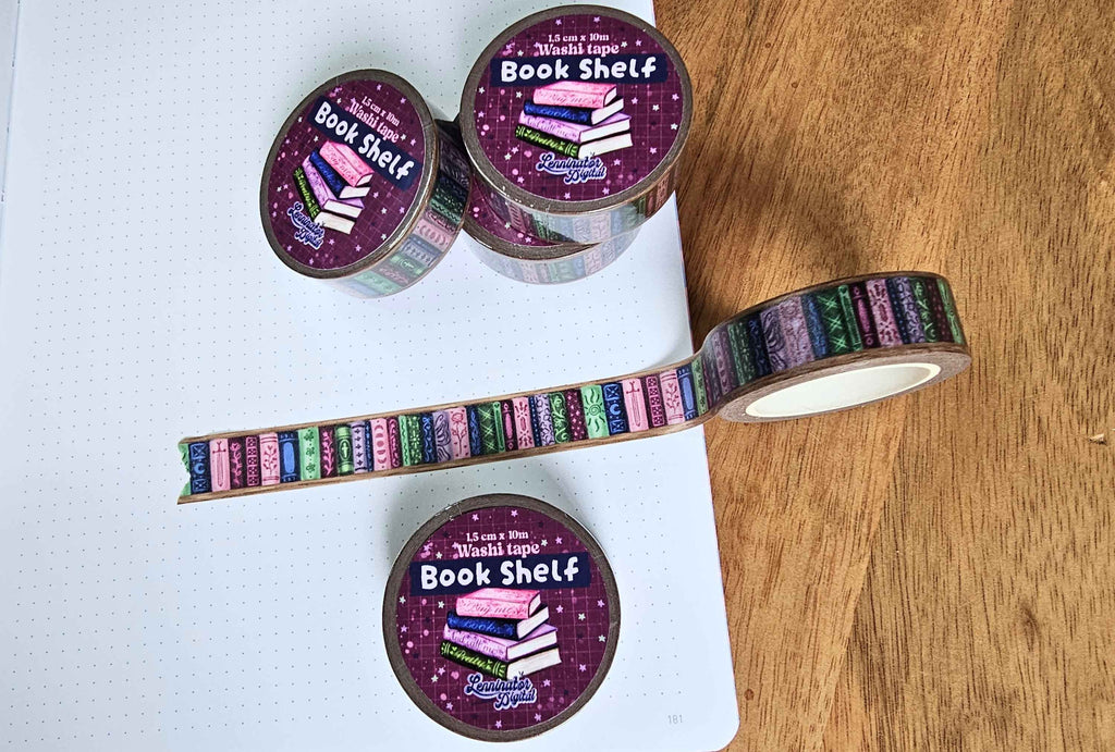 A roll of decorative washi tape with a colorful book spine pattern is displayed on a white grid notebook and a wooden surface. The tape features a purple starry background with pink, blue, green, and orange book spines.