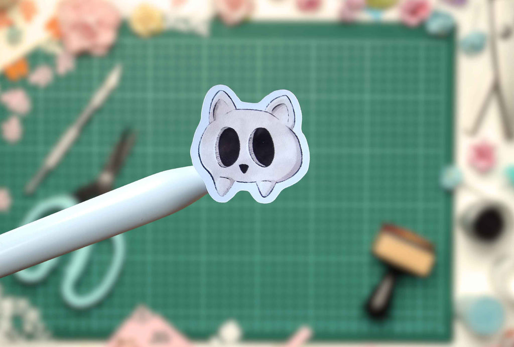 A small die-cut sticker of a white cartoon cat face with large black eyes is held on a white stylus over a green cutting mat with craft supplies in the background.