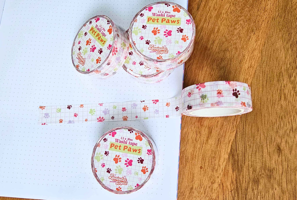 Several rolls of decorative washi tape with a colorful 'It's a World Tape Pet Paws' pattern are arranged on a white dotted notebook and a wooden surface. The tape features small paw prints in pink, orange, green, and brown over a light grid background.